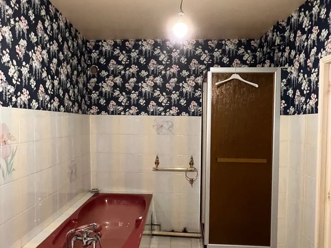 Salle de bain d'époque : papier peint à fleurs sur faïence blanche, baignoire bordeaux, cabine de douche en verre fumé, ampoule nue au plafond.